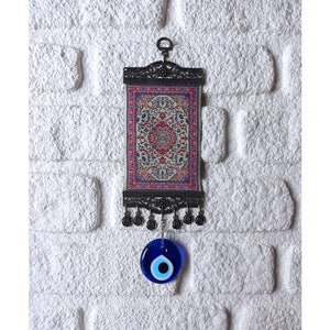 May include: A small tapestry with a traditional Turkish rug design hangs from a metal frame with a blue evil eye charm hanging below. The tapestry is framed with a black metal frame and has tassels hanging from the bottom.