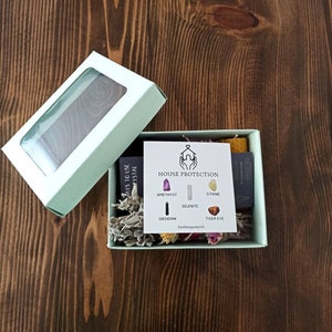May include: A light green box with a clear window lid, containing a small white card with the text "HOUSE PROTECTION" and images of crystals. The box also contains dried flowers and sage.