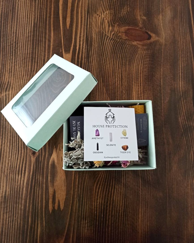 May include: A light green box with a clear window lid, containing a small white card with the text "HOUSE PROTECTION" and images of crystals. The box also contains dried flowers and sage.