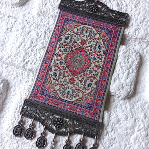 May include: A small tapestry with a traditional Persian rug design in shades of red, blue, and cream. The tapestry is framed with a silver metal frame and has a silver chain hanging from the bottom.