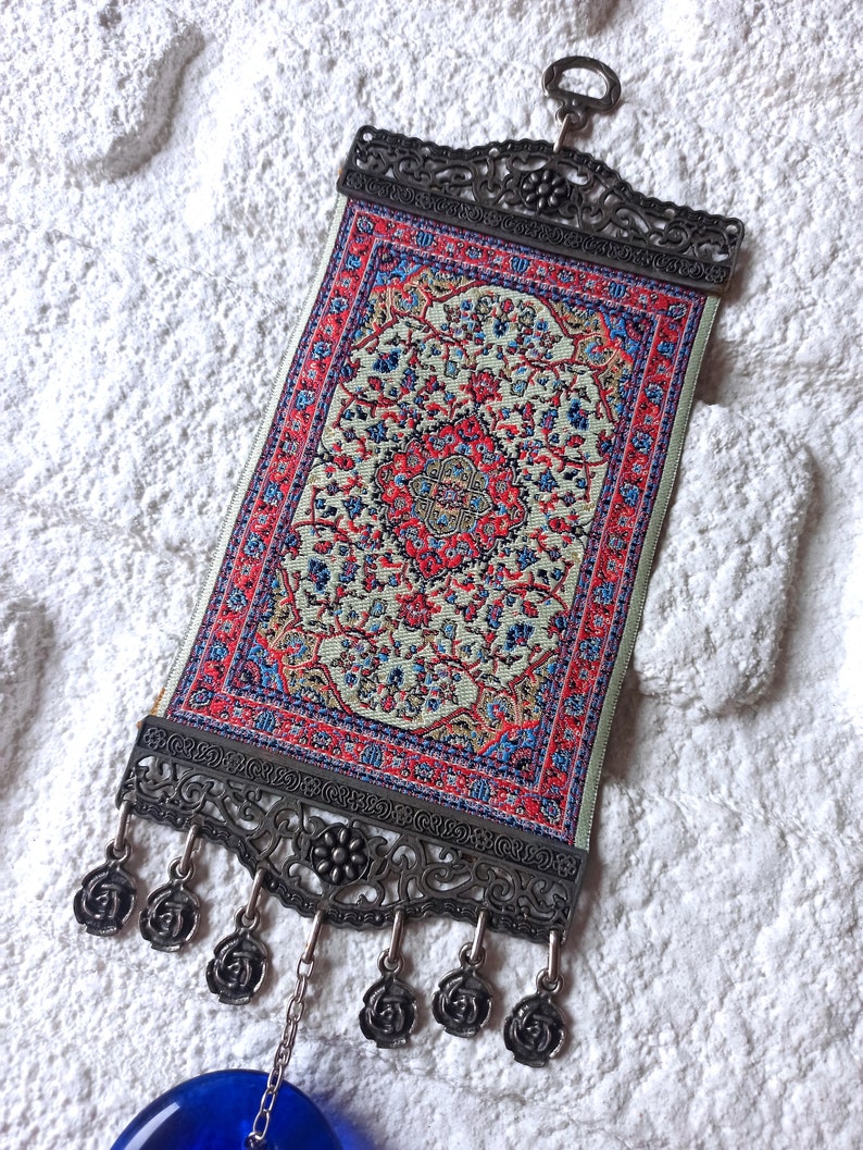 May include: A small tapestry with a traditional Persian rug design in shades of red, blue, and cream. The tapestry is framed with a silver metal frame and has a silver chain hanging from the bottom.