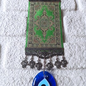 May include: A green and gold patterned fabric wall hanging with a decorative metal frame and a blue evil eye charm hanging from the bottom.