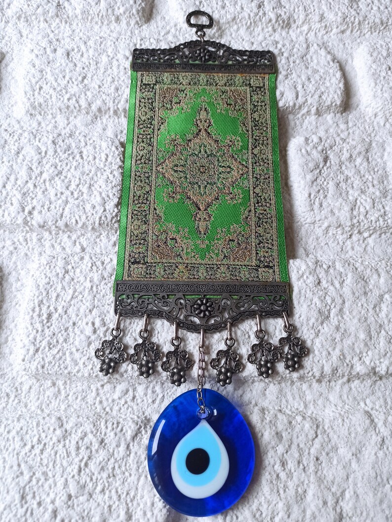 May include: A green and gold patterned fabric wall hanging with a decorative metal frame and a blue evil eye charm hanging from the bottom.