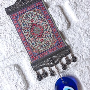 May include: A small tapestry with a traditional Turkish rug design hangs from a silver metal frame with a blue evil eye charm hanging below.