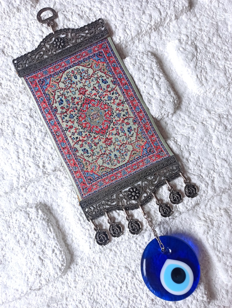 May include: A small tapestry with a traditional Turkish rug design hangs from a silver metal frame with a blue evil eye charm hanging below.