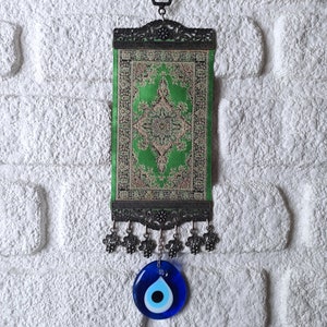 May include: A green and gold patterned fabric wall hanging with a silver frame and a blue evil eye charm hanging from the bottom.