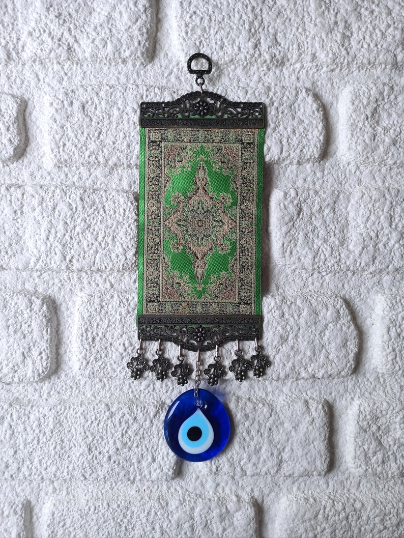 May include: A green and gold patterned fabric wall hanging with a silver frame and a blue evil eye charm hanging from the bottom.