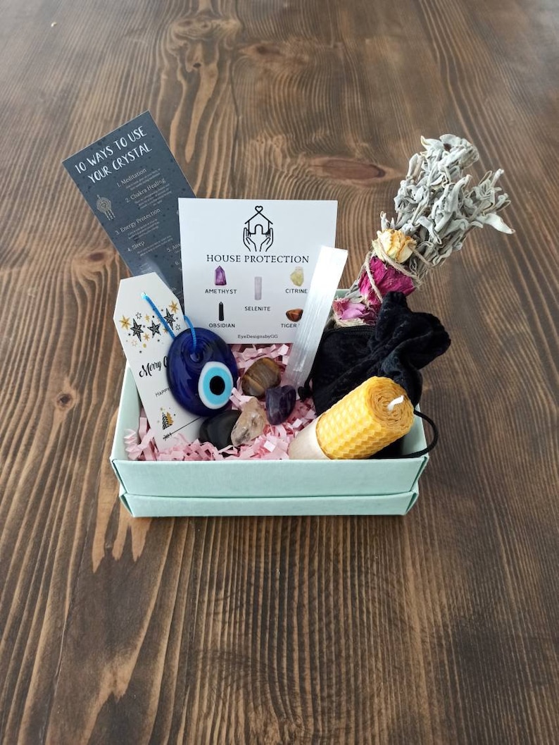May include: A light blue box filled with crystals, a sage bundle, a yellow candle, and a blue evil eye charm. The box is labelled "House Protection" and lists the crystals included: amethyst, citrine, selenite, obsidian, and tiger's eye.