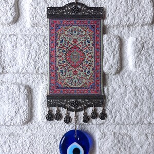 May include: A small tapestry with a traditional Turkish rug design hangs from a metal frame with a blue evil eye charm hanging below.