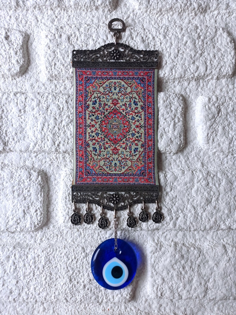 May include: A small tapestry with a traditional Turkish rug design hangs from a metal frame with a blue evil eye charm hanging below.