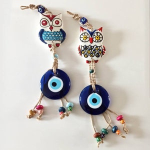 May include: Two ceramic owl charms with blue and white painted details, each with a blue evil eye charm hanging below. The owls have red, yellow, and black accents. The charms are strung on twine with brown beads.