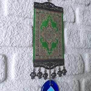 May include: A green and gold tapestry with an intricate design hangs from a metal hook. It has a blue evil eye charm hanging from the bottom.