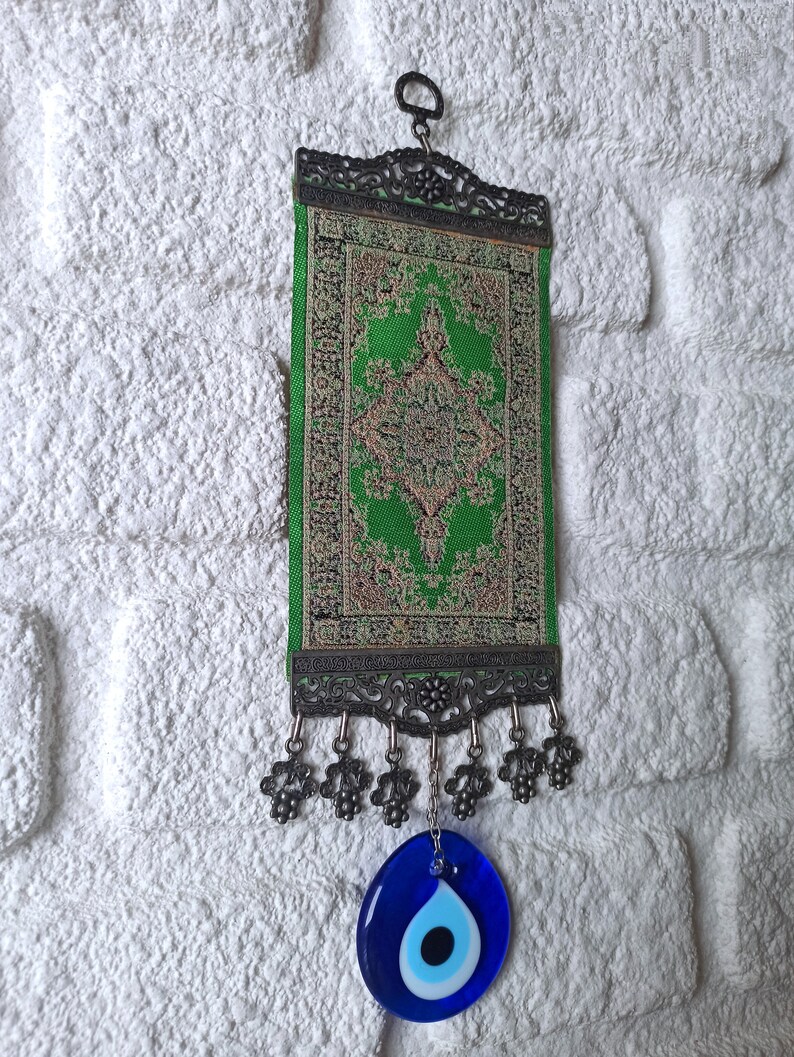 May include: A green and gold tapestry with an intricate design hangs from a metal hook. It has a blue evil eye charm hanging from the bottom.