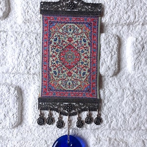 May include: A small tapestry with a traditional Turkish rug design hangs from a metal frame with a blue evil eye charm hanging below.