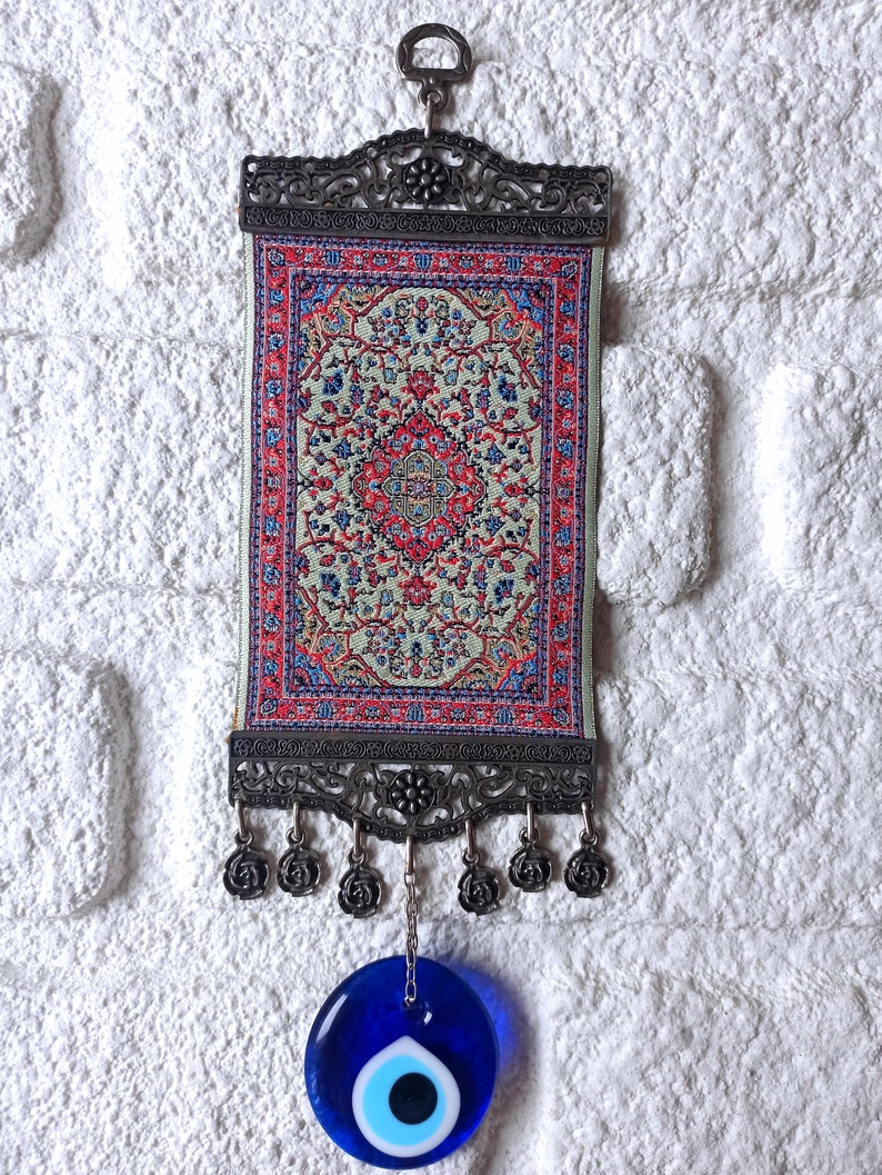May include: A small tapestry with a traditional Turkish rug design hangs from a metal frame with a blue evil eye charm hanging below.