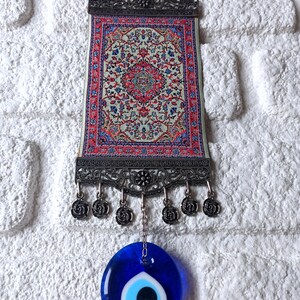 May include: A small tapestry with a traditional Turkish rug design hangs from a metal frame with a blue evil eye charm hanging from the bottom.