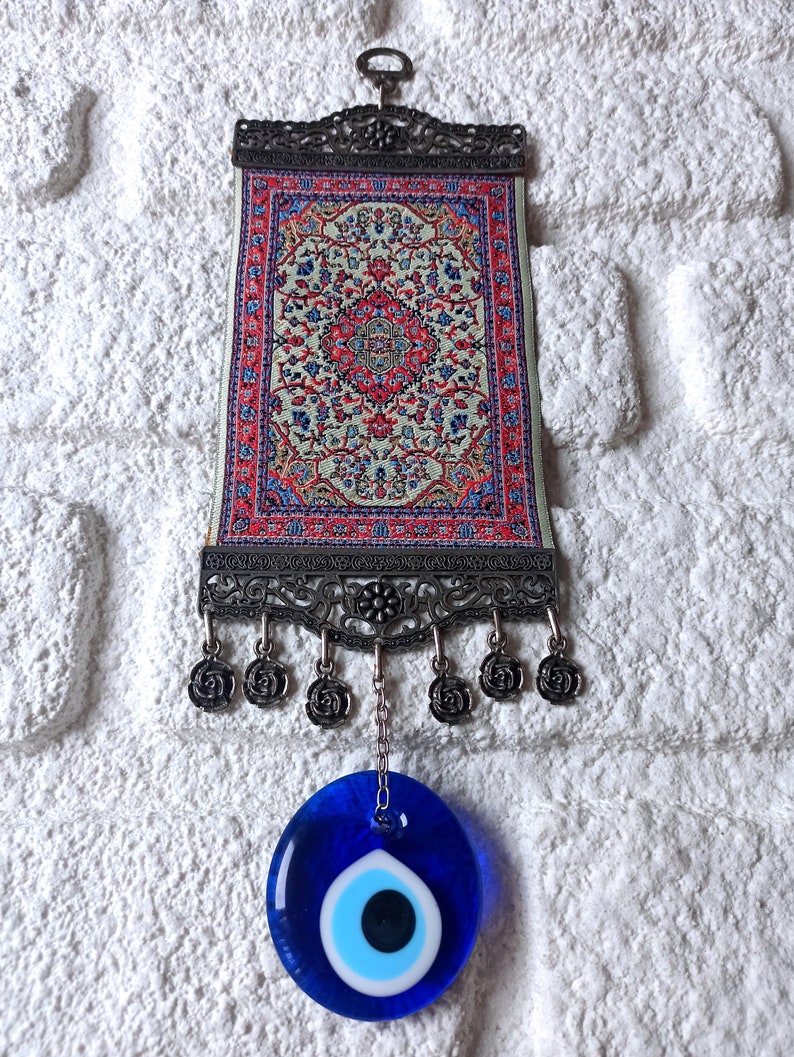 May include: A small tapestry with a traditional Turkish rug design hangs from a metal frame with a blue evil eye charm hanging from the bottom.