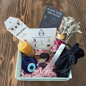 May include: A light blue gift box filled with crystals, a candle, a sage smudge stick, a small pouch, and a blue evil eye charm. The box is decorated with pink tissue paper. The box also includes a card that says "House Protection" and lists different crystals, including amethyst, citrine, selenite, obsidian, and tiger's eye. The card also includes a card that says "10 Ways to Use Your Crystal" and lists different ways to use crystals, including meditation, chakra healing, energy protection, and sleep.