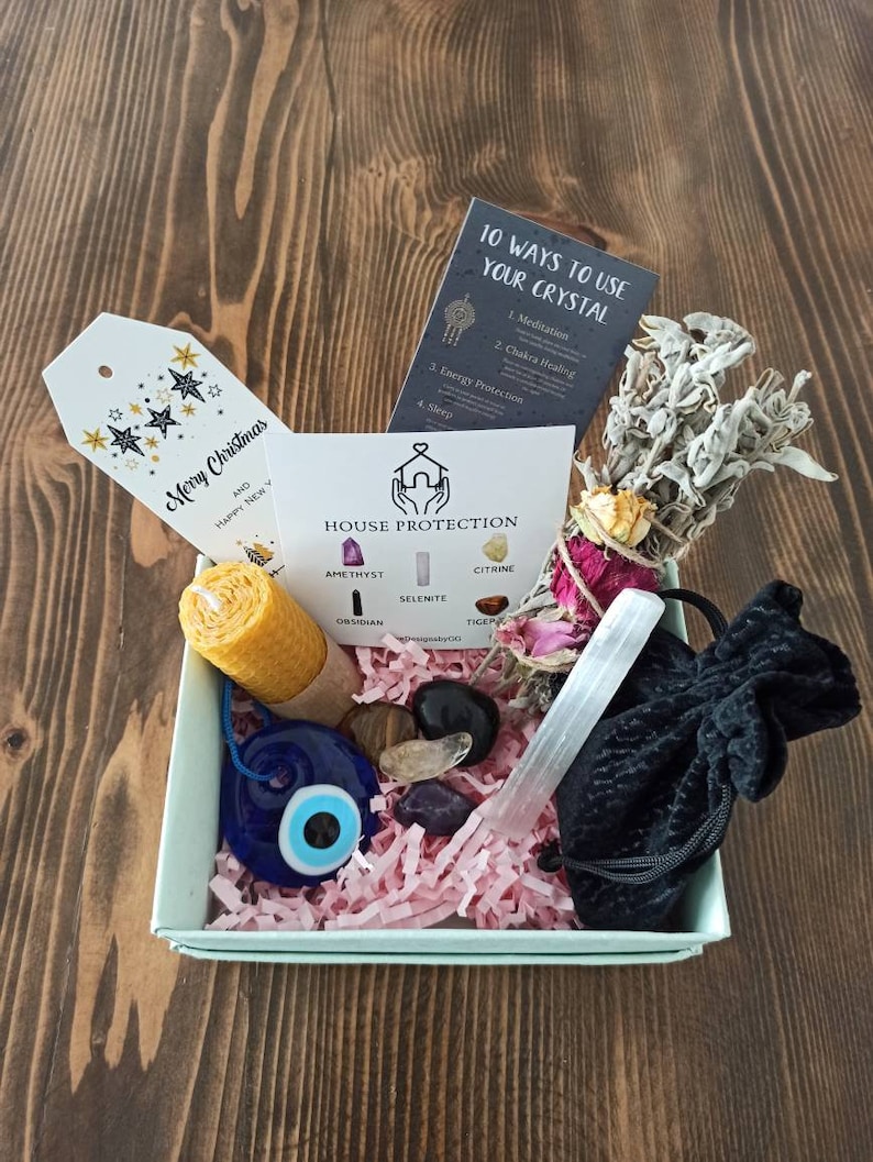 May include: A light blue gift box filled with crystals, a candle, a sage smudge stick, a small pouch, and a blue evil eye charm. The box is decorated with pink tissue paper. The box also includes a card that says "House Protection" and lists different crystals, including amethyst, citrine, selenite, obsidian, and tiger's eye. The card also includes a card that says "10 Ways to Use Your Crystal" and lists different ways to use crystals, including meditation, chakra healing, energy protection, and sleep.