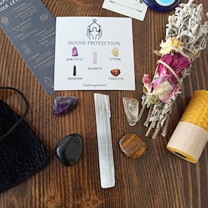 May include: A set of crystals for house protection, including amethyst, selenite, citrine, obsidian, and tiger's eye. The crystals are arranged on a wooden surface with a white card that says "House Protection" and lists the names of the crystals. There is also a sage smudge stick, a white candle, and a small pouch.