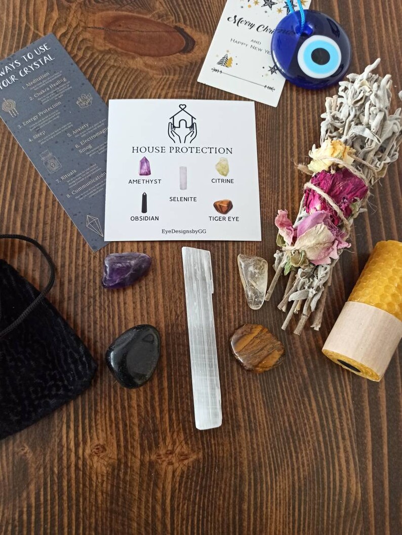 May include: A set of crystals for house protection, including amethyst, selenite, citrine, obsidian, and tiger's eye. The crystals are arranged on a wooden surface with a white card that says "House Protection" and lists the names of the crystals. There is also a sage smudge stick, a white candle, and a small pouch.