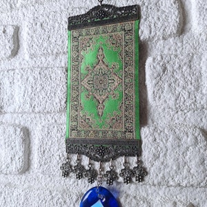 May include: A green and gold patterned fabric wall hanging with a silver frame and a blue evil eye charm hanging from the bottom.