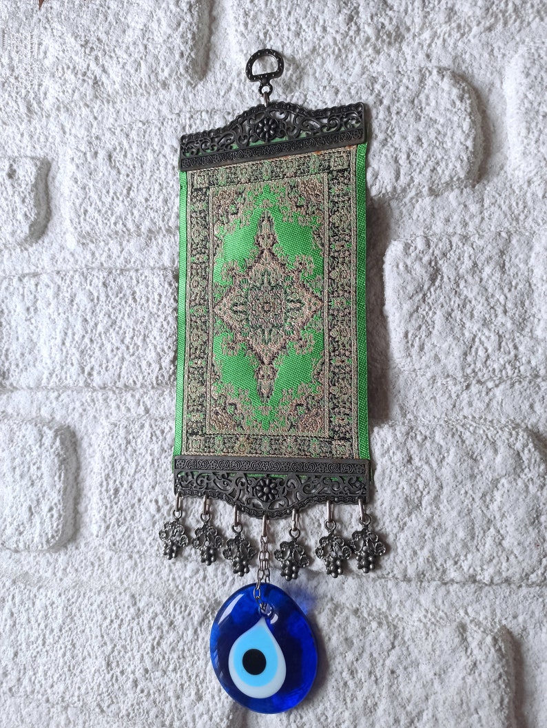 May include: A green and gold patterned fabric wall hanging with a silver frame and a blue evil eye charm hanging from the bottom.