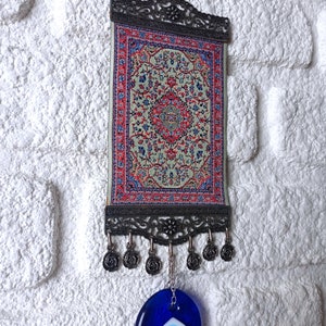 May include: A small tapestry with a traditional Turkish rug design hangs from a metal frame with a blue evil eye charm hanging below.