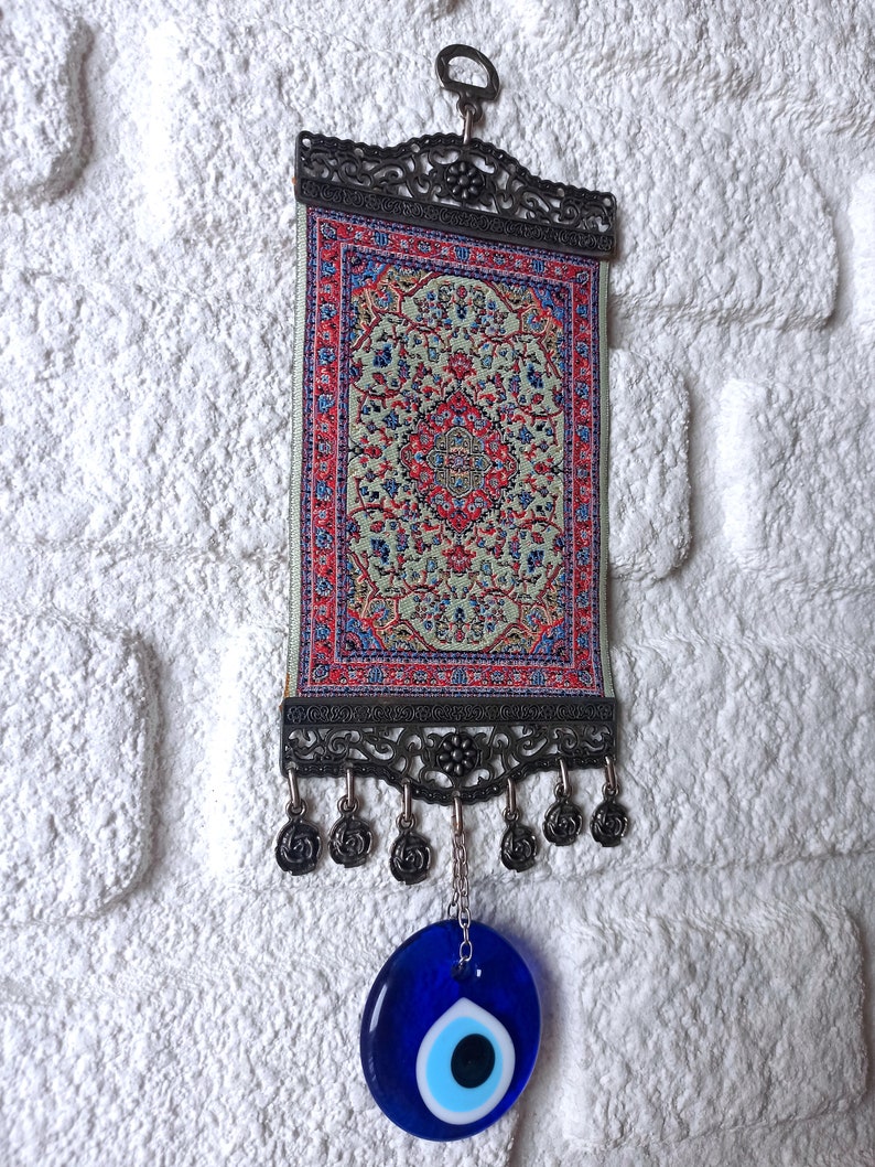 May include: A small tapestry with a traditional Turkish rug design hangs from a metal frame with a blue evil eye charm hanging below.