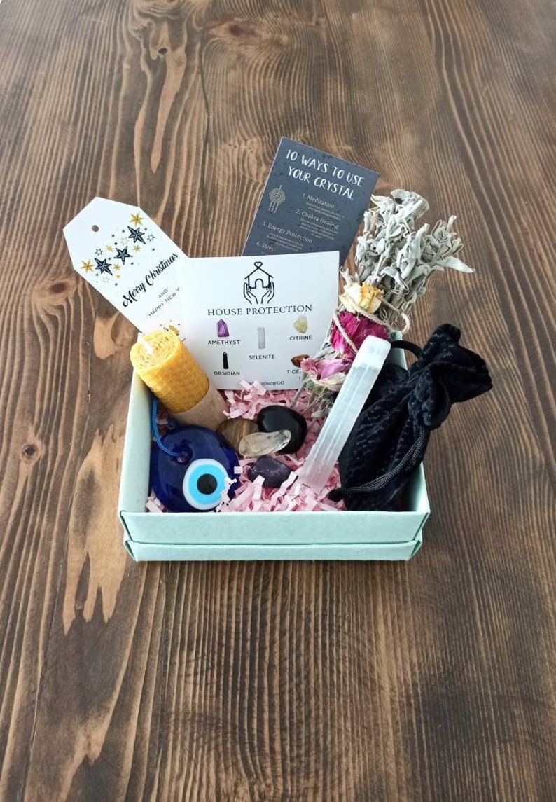 May include: A light blue gift box filled with crystals, a candle, sage, and an evil eye charm. The box also includes a card that says "House Protection" and lists the crystals included: amethyst, citrine, selenite, obsidian, and tiger's eye.