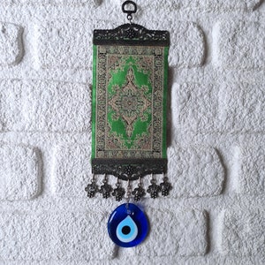 May include: A green and gold tapestry with a traditional design hangs from a metal hook. A blue evil eye charm hangs from the bottom of the tapestry.