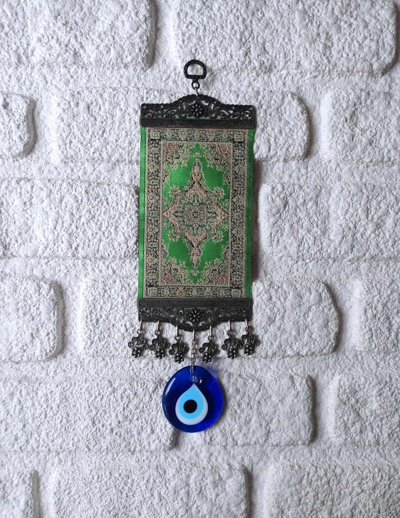 May include: A green and gold tapestry with a traditional design hangs from a metal hook. A blue evil eye charm hangs from the bottom of the tapestry.