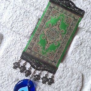 May include: A green and gold tapestry with a decorative metal frame and a blue evil eye charm hanging from it. The tapestry has a tassel fringe and is hung on a white wall.