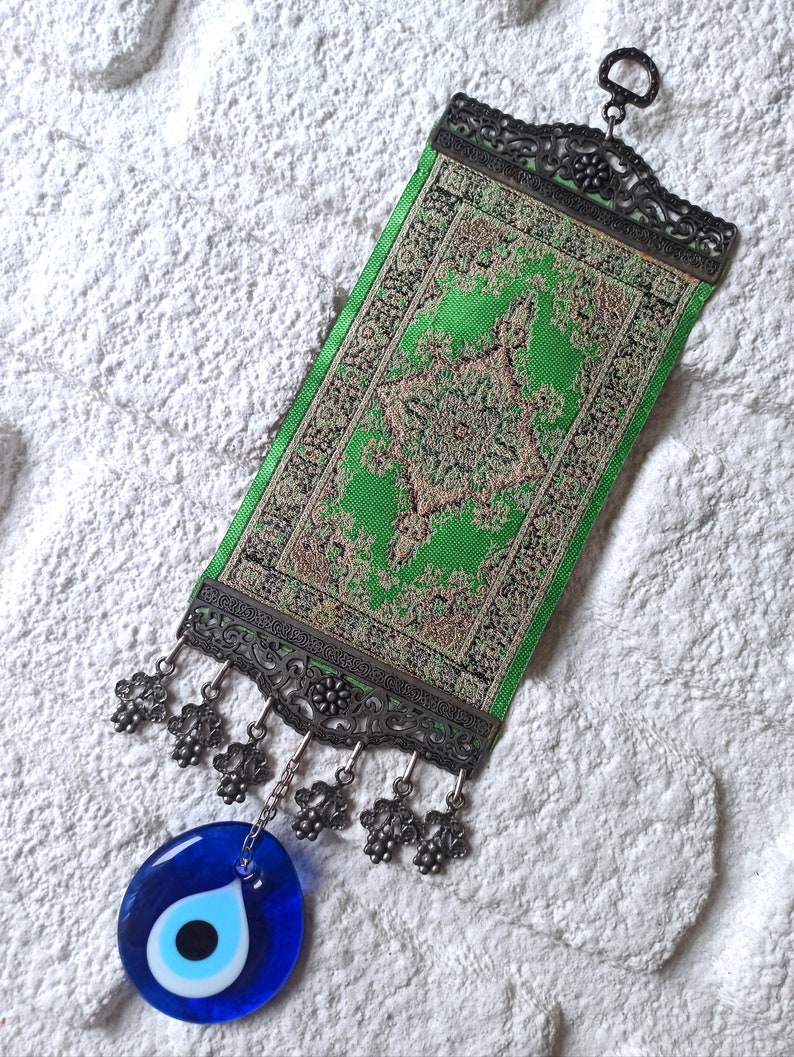 May include: A green and gold tapestry with a decorative metal frame and a blue evil eye charm hanging from it. The tapestry has a tassel fringe and is hung on a white wall.
