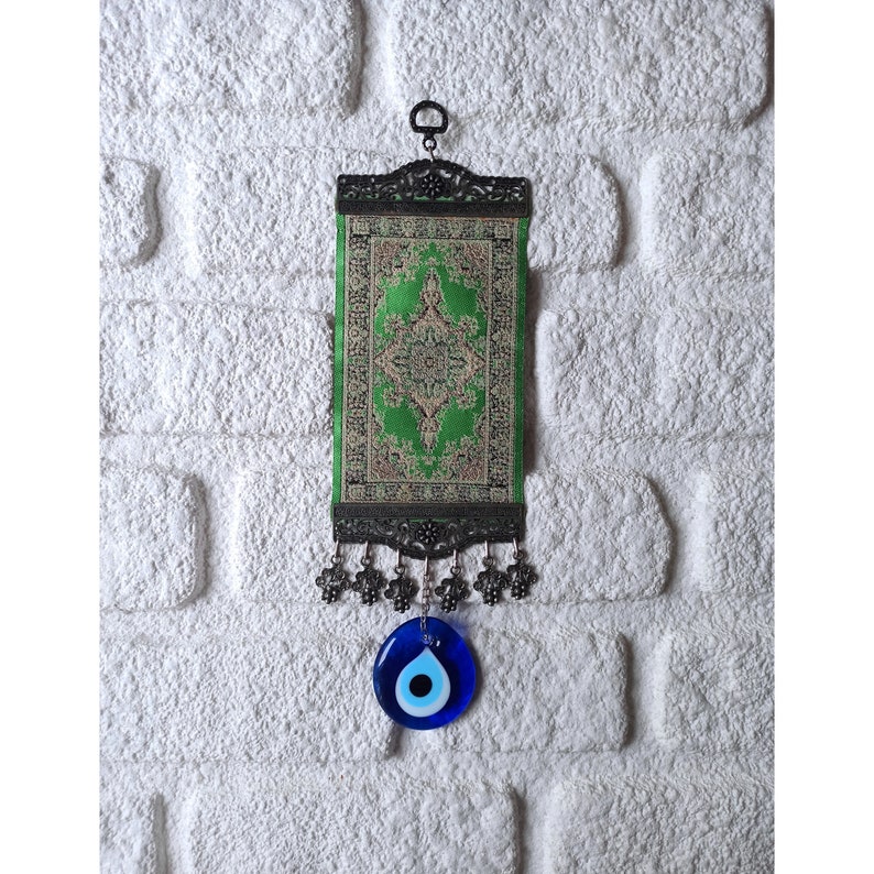 May include: A green and gold patterned fabric wall hanging with a black metal frame and tassels. A blue evil eye charm hangs from the bottom of the frame.