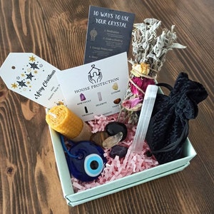 May include: A light blue box filled with pink crinkle paper and crystals, a white sage smudge stick, a small candle, a black velvet bag, a white selenite wand, and a blue evil eye charm. The box also includes a card with the text "House Protection" and a list of crystals: Amethyst, Selenite, Citrine, Obsidian, Tiger.