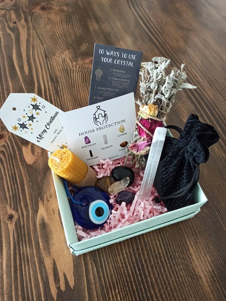 May include: A light blue box filled with pink crinkle paper and crystals, a white sage smudge stick, a small candle, a black velvet bag, a white selenite wand, and a blue evil eye charm. The box also includes a card with the text "House Protection" and a list of crystals: Amethyst, Selenite, Citrine, Obsidian, Tiger.