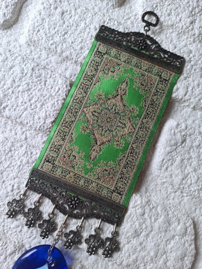May include: A green and gold patterned fabric wall hanging with a metal frame and tassels. The hanging features a blue glass ornament.