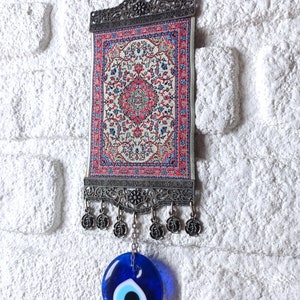 May include: A small tapestry with a traditional Turkish rug design hangs from a metal frame with a blue evil eye charm hanging below.