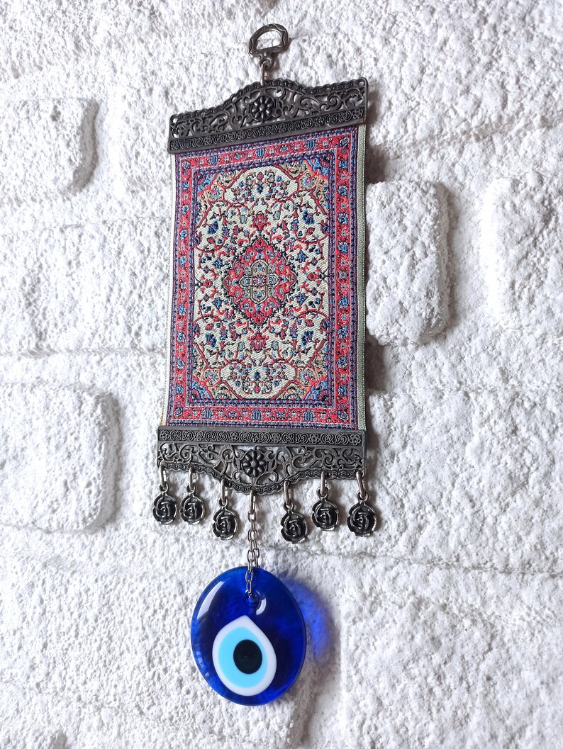 May include: A small tapestry with a traditional Turkish rug design hangs from a metal frame with a blue evil eye charm hanging below.