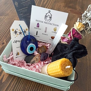 May include: A gift box filled with crystals, a candle, and a sage smudge stick. The box is decorated with pink tissue paper and a card that says "House Protection". The crystals are amethyst, selenite, citrine, obsidian, tiger's eye, and a blue evil eye charm.