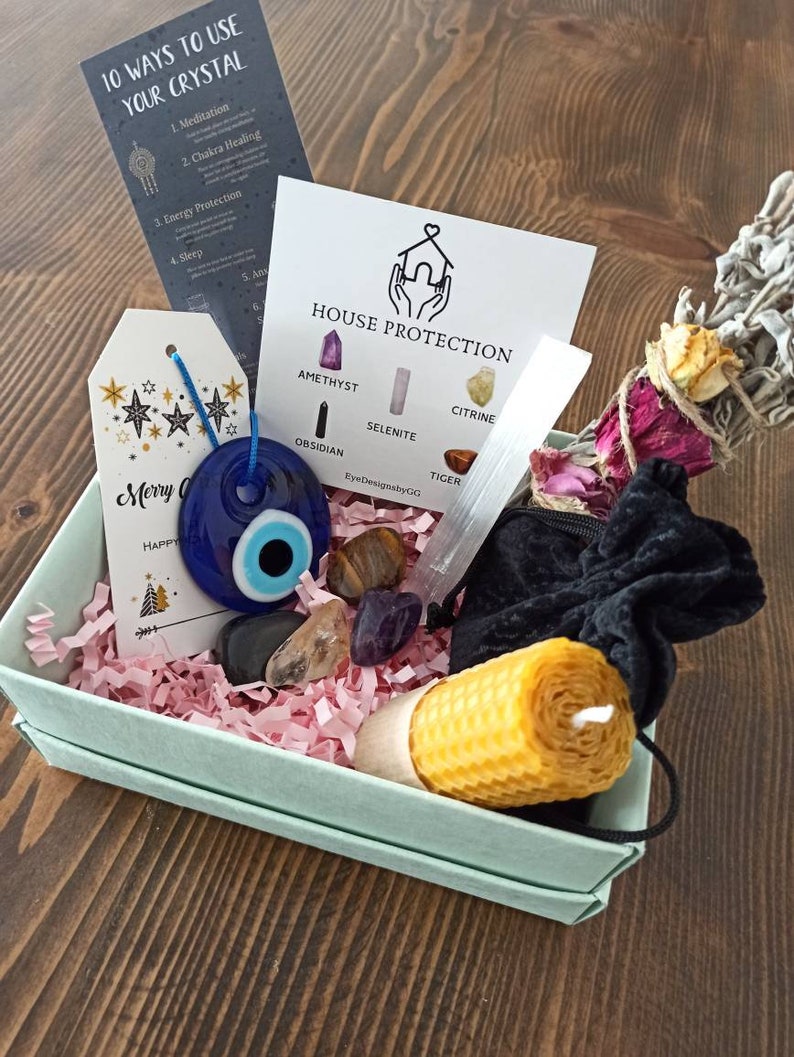 May include: A gift box filled with crystals, a candle, and a sage smudge stick. The box is decorated with pink tissue paper and a card that says "House Protection". The crystals are amethyst, selenite, citrine, obsidian, tiger's eye, and a blue evil eye charm.