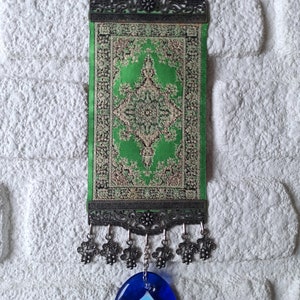 May include: A green and gold patterned fabric wall hanging with a blue evil eye charm. The hanging has a metal frame with decorative details and tassels.