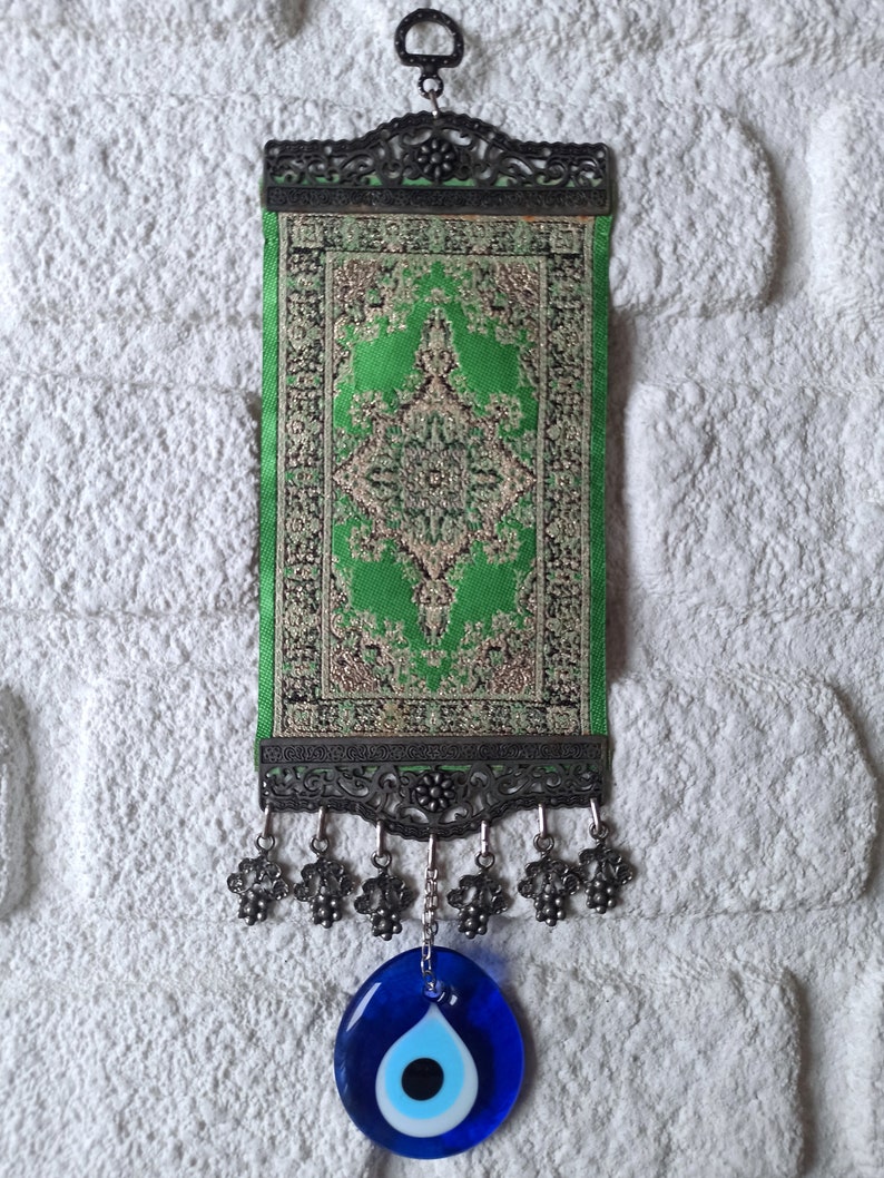 May include: A green and gold patterned fabric wall hanging with a blue evil eye charm. The hanging has a metal frame with decorative details and tassels.