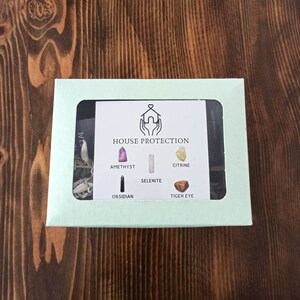 May include: A light green box with a clear window showing a set of crystals for house protection. The crystals are amethyst, selenite, citrine, obsidian, and tiger's eye. The box has the text "HOUSE PROTECTION" printed on it.