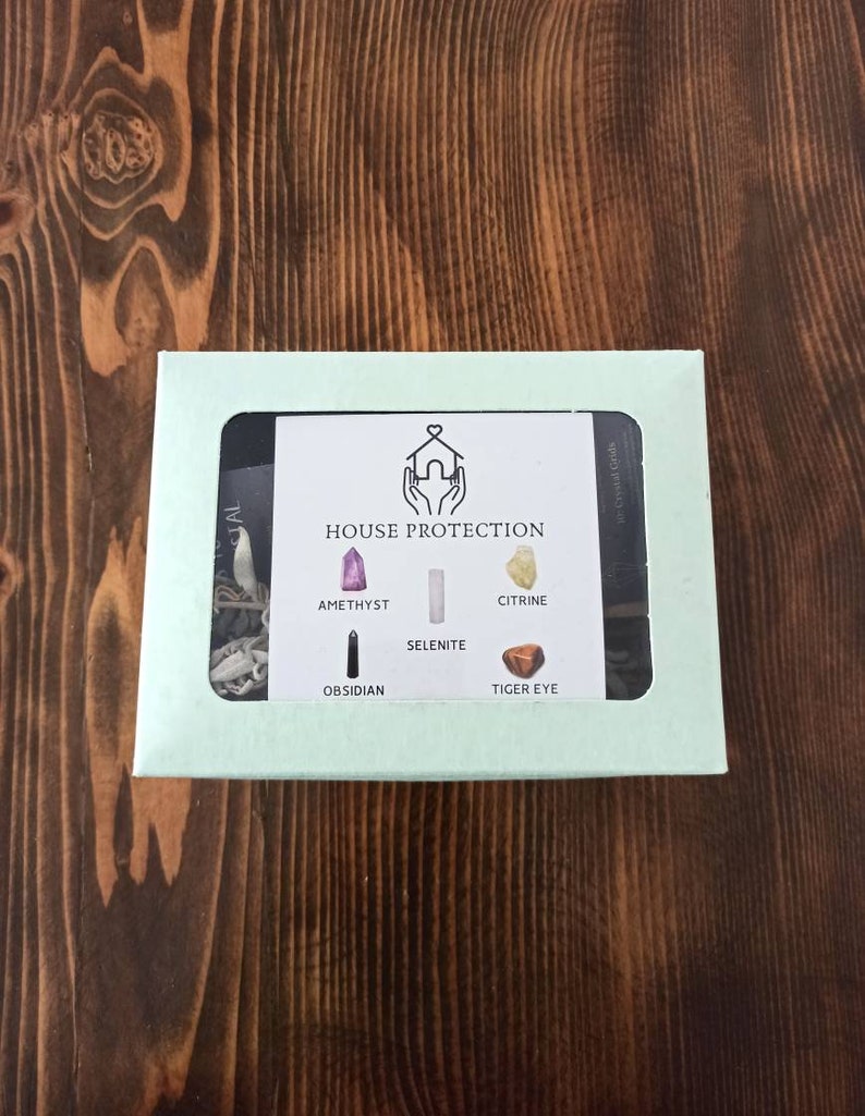 May include: A light green box with a clear window showing a set of crystals for house protection. The crystals are amethyst, selenite, citrine, obsidian, and tiger's eye. The box has the text "HOUSE PROTECTION" printed on it.