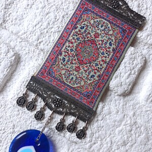 May include: A small, decorative wall hanging featuring a traditional Turkish rug design with intricate floral patterns in shades of red, blue, and green. The rug is framed by a silver metal frame with a hanging loop and decorative tassels. A blue evil eye charm hangs from the bottom of the frame.