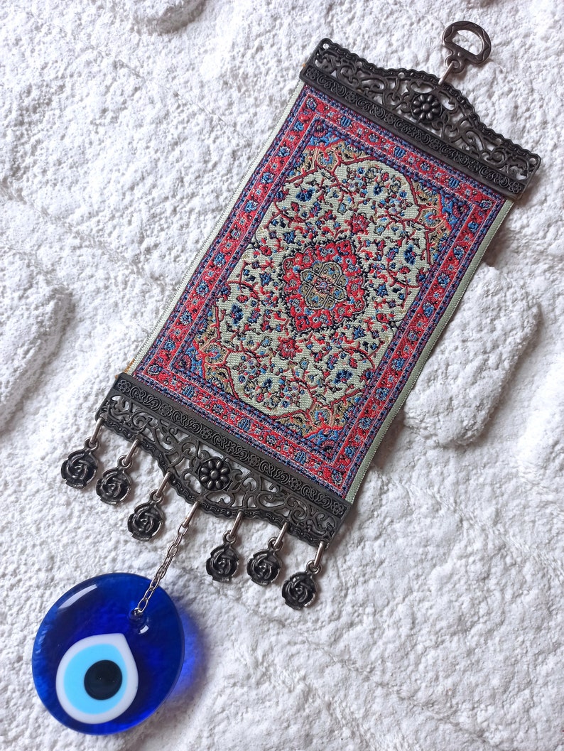 May include: A small, decorative wall hanging featuring a traditional Turkish rug design with intricate floral patterns in shades of red, blue, and green. The rug is framed by a silver metal frame with a hanging loop and decorative tassels. A blue evil eye charm hangs from the bottom of the frame.