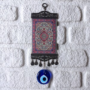 May include: A small tapestry with a traditional Turkish rug design hangs from a metal frame with a blue evil eye charm hanging below.