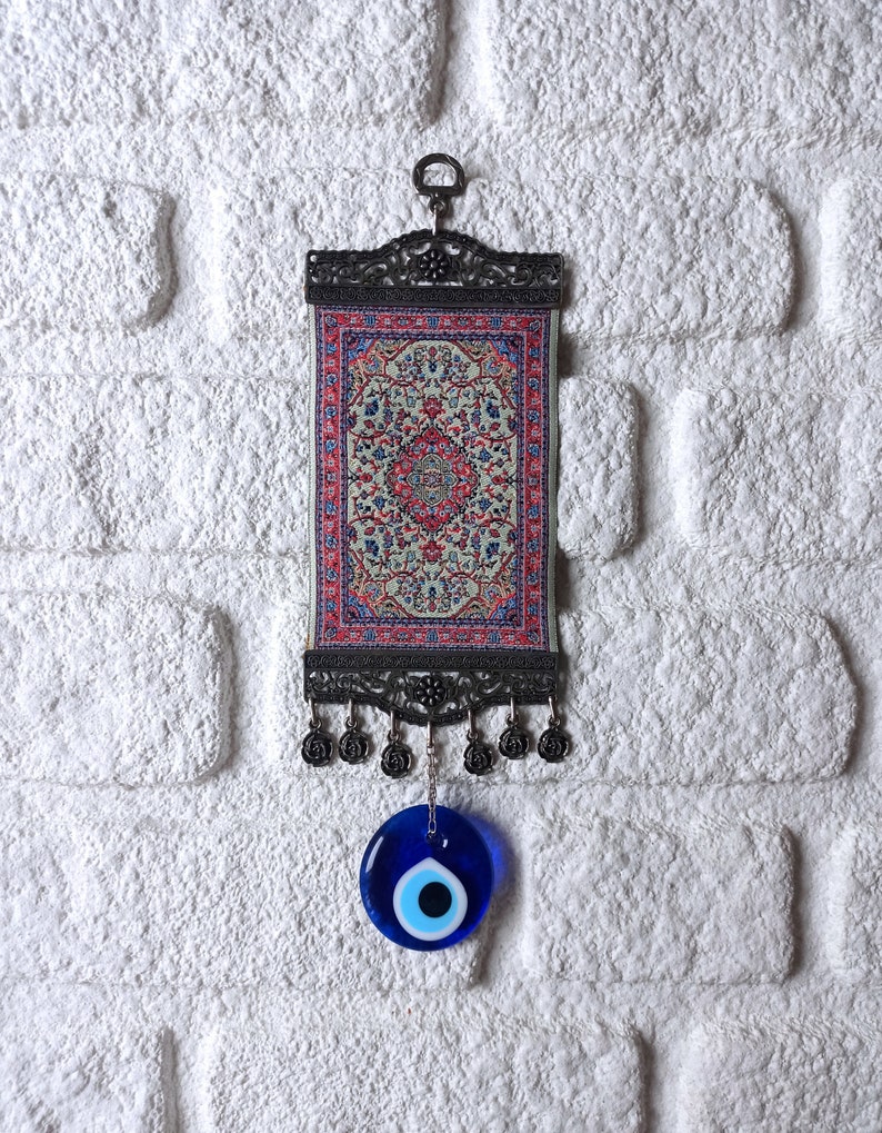 May include: A small tapestry with a traditional Turkish rug design hangs from a metal frame with a blue evil eye charm hanging below.